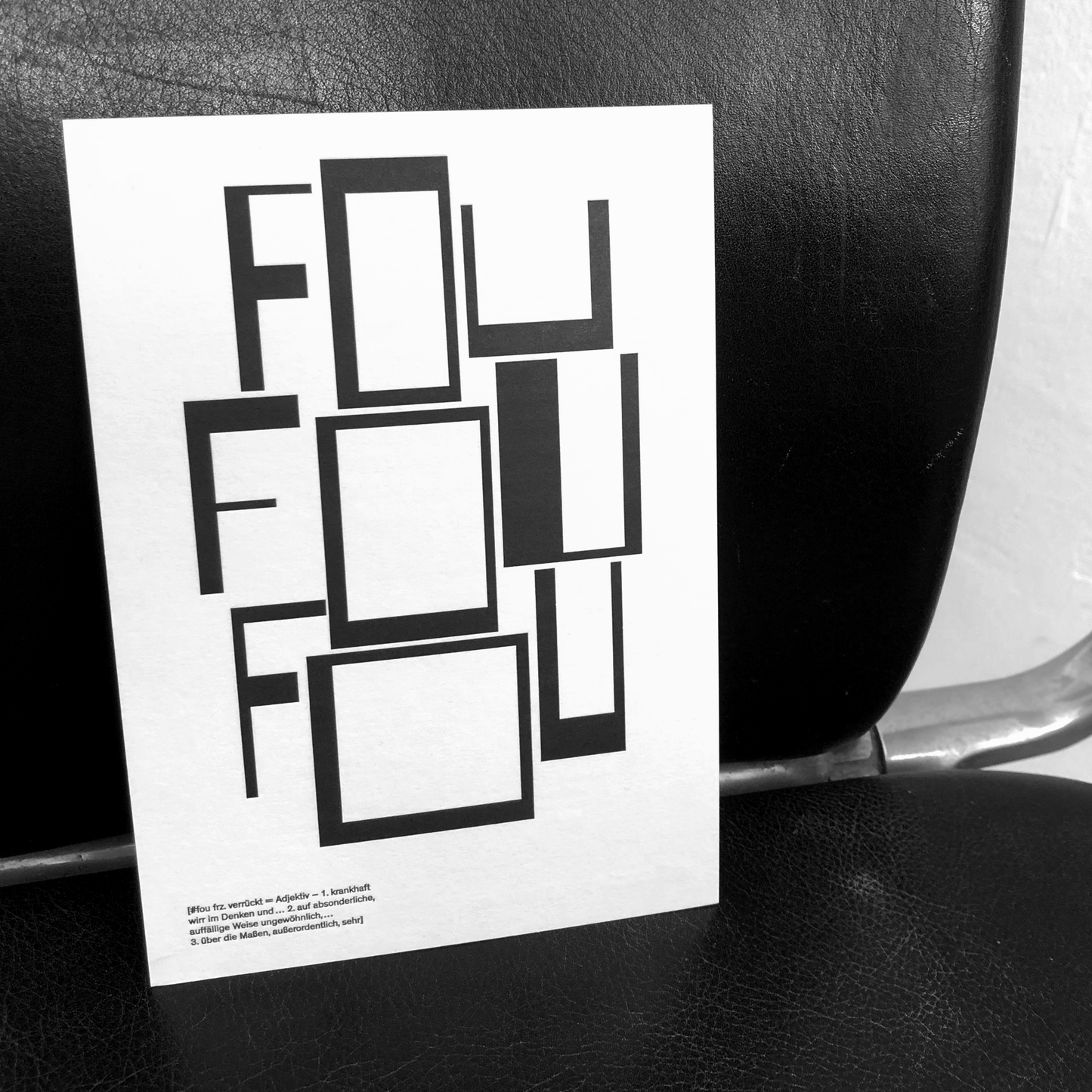 Schwarz-weißes Foto: Design mit Wort "Fou" auf Papier, schwarzer Lederstuhl im Hintergrund.